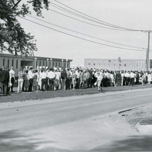 Crowd of students lined up on sidewalk and lawn to enter flat-topped one-story building