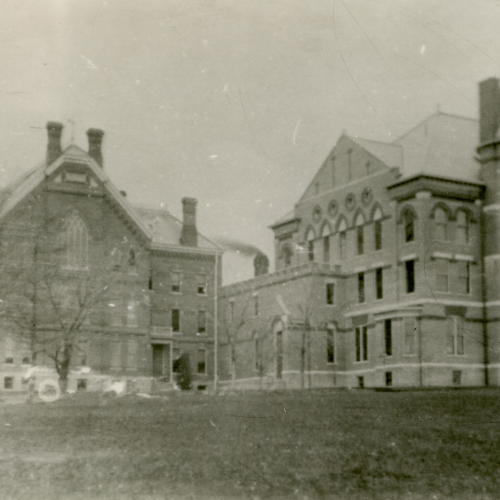 Three and a half story brick building on left connected with two-story passageway to taller three and a half story building on right, smokestack in background 