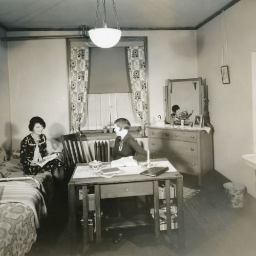 One woman sitting at a desk in the middle of a dorm room and another woman sitting on a bed to the left; there is a window at the back of the room, a dresser to the back right, and a sink on the right wall