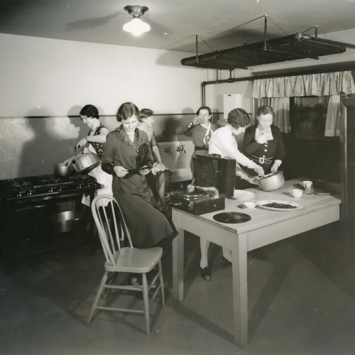 Six women in a kitchen, some making food and one putting a record in the record player on the central table