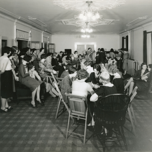 Group of women in a lounge, playing in a band in the back right corner, seated around three or four tables in the middle of the room, some playing cards, and standing and sitting along the walls