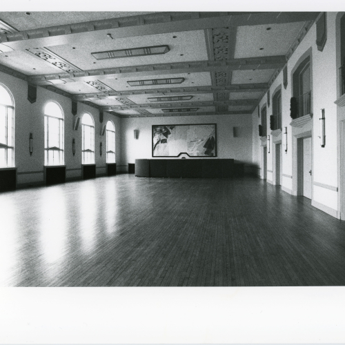 Empty room with wooden floor, archways and balconies on right, windows on left, and artwork on far wall