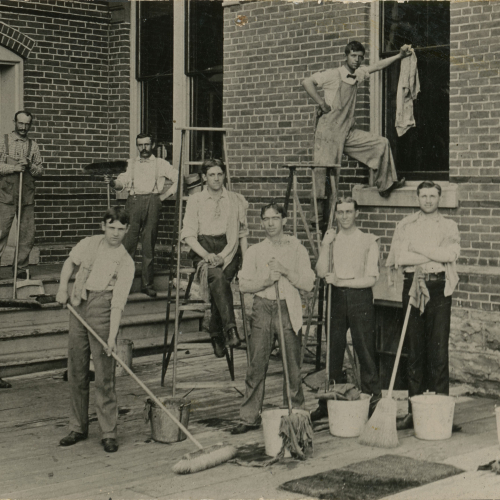 10 men with mops, buckets, and other cleaning supplies on a wooden porch outside a brick building