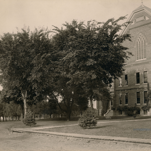 Wooden walkway, gravel road, and tall trees outside a three and a half story brick building with pointed arched window on upper floor