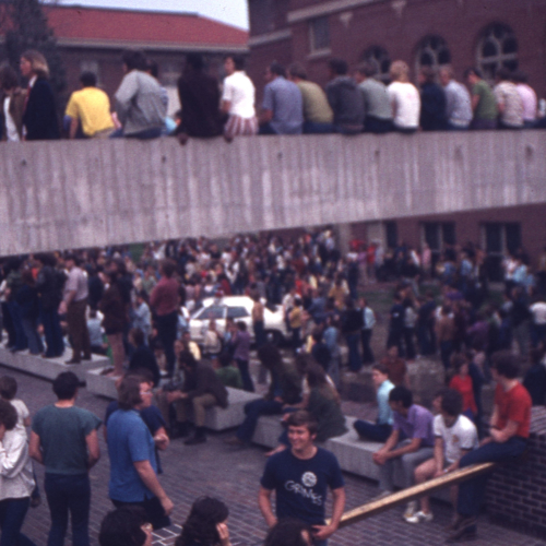 Crowd of people sitting/standing on a concrete bridge as well as a rooftop level under it and a plaza at ground level