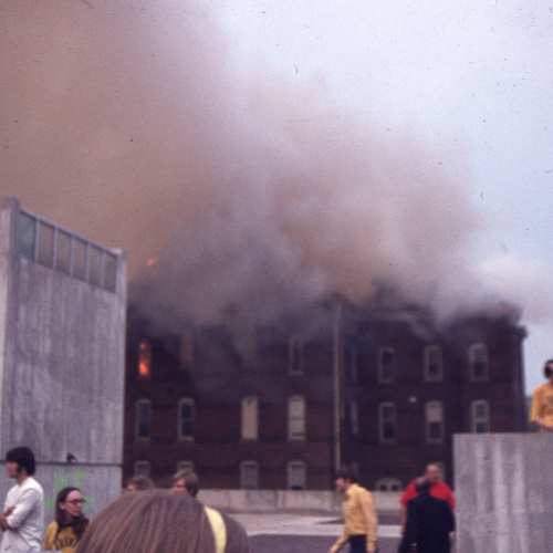 A brick building on fire, some flames to the left but smoke obscuring most of it, and people watching from the flat rooftop of a nearby building