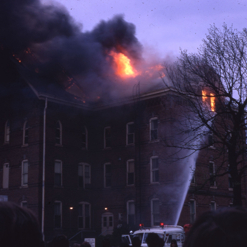 Fire truck spraying water at flames and smoke engulfing top of a three and a half story brick building