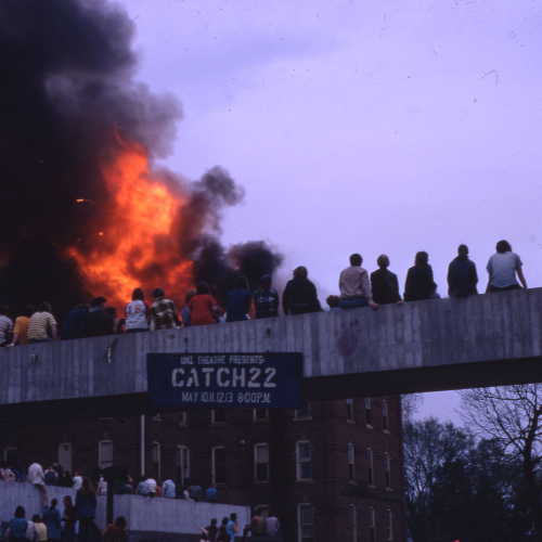 People seated on an elevated concrete walkway as well as the rooftop below it, watching a nearby brick building engulfed with flames and smoke