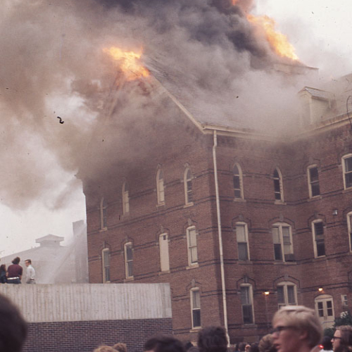 Crowd watching a three and a half story brick building, which is on fire near the roof