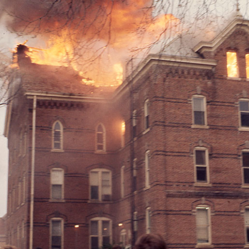 A red brick three and a half story building with flames engulfing the roof and visible through the attic window
