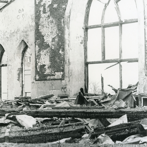 Burned wood and other debris fallen to the floor of a room with a busted-out large pointed arched window