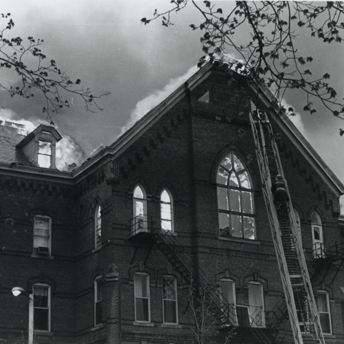 Firefighter climbing a ladder propped against the facade of a brick building with pointed arched window near the top; building has flames visible through the window and engulfing the roof