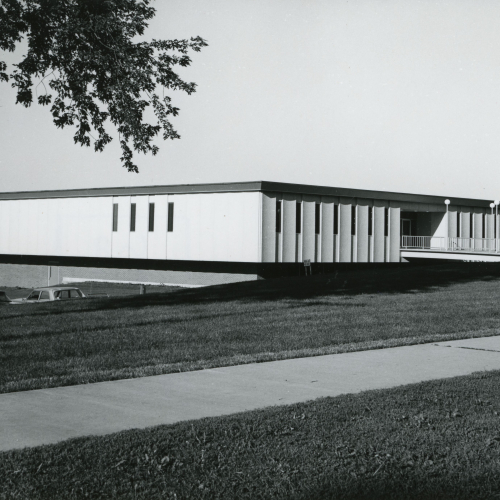 Flat-topped one-story building with light-colored sides and darker roof and lower concrete level opened to parking lot
