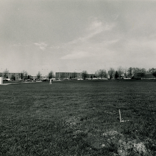 Open lawn space with flat-topped building and smokestack to right, dorm buildings ahead, and welcome pillars to left