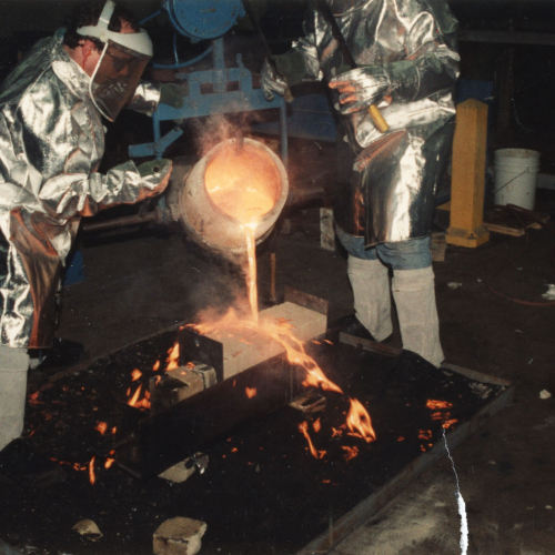 Two people in protective suits and helmets pouring molten metal into a mold