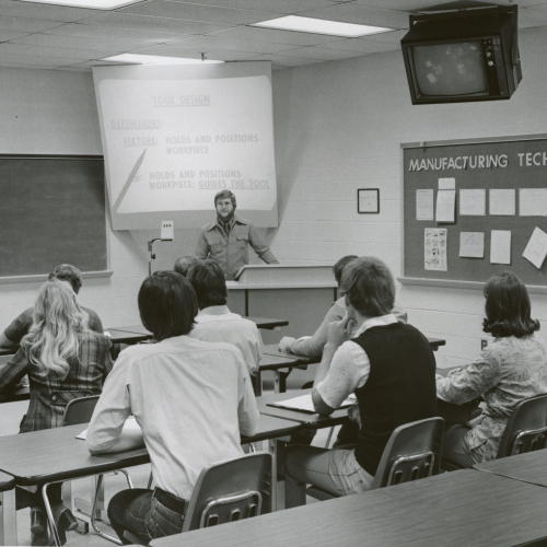 Instructor presenting slideshow to 8 students in classroom; title of bulletin board to the right is Manufacturing Technology