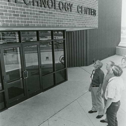 Three men look up at the entrance to a building with partially visible lettering reading Technology Center