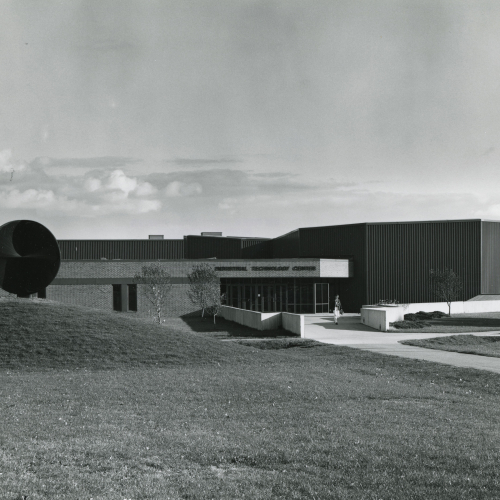 Building with entrance in center and brick wing to the left, metal wing to the right, across a lawn with a hill and circular sculpture/dish at its top