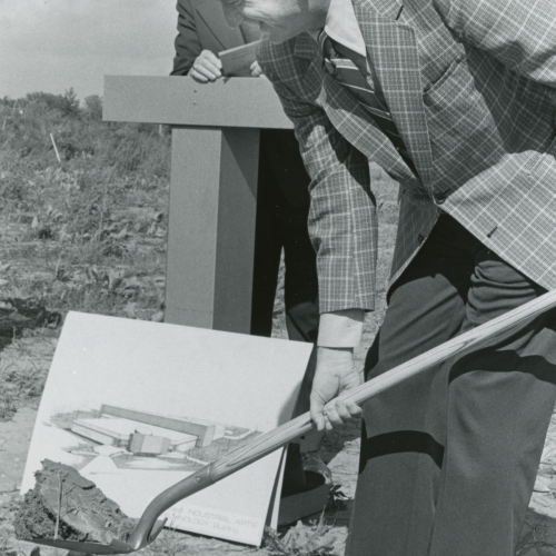 In foreground, man turning shovelful of dirt; another man leans on podium behind him with architectural rendering propped in front
