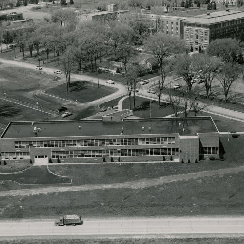 Aerial of a two-story flat-roofed building with many windows along the side, other campus buildings across a tree-lined street in the background