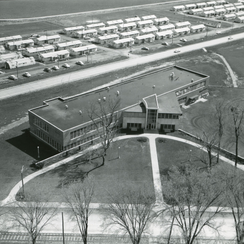 Aerial of a two-story flat-roofed building with many windows; in background, quonset huts and metal buildings composing Sunset Village