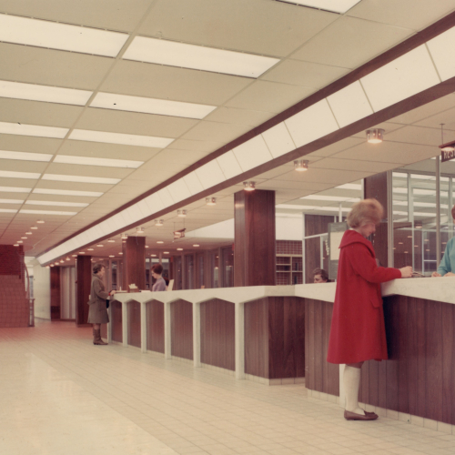 A woman helping a patron at the reserve desk in the foreground, and another woman helping another patron at the check out desk in background