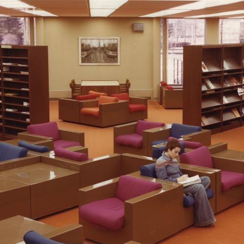 One student seated in one of lounge chairs in a grouping and another browsing periodical shelves