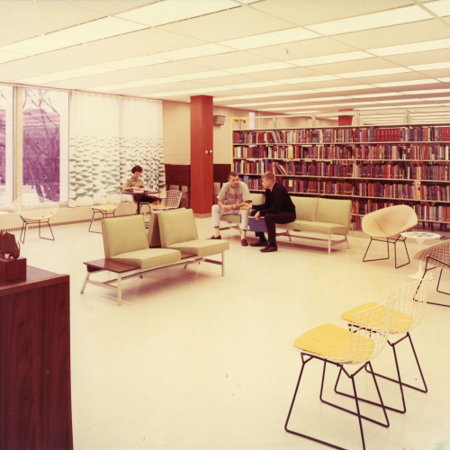 Two students talking on a sofa and one reading at a table in a lounge area with library stacks in the background