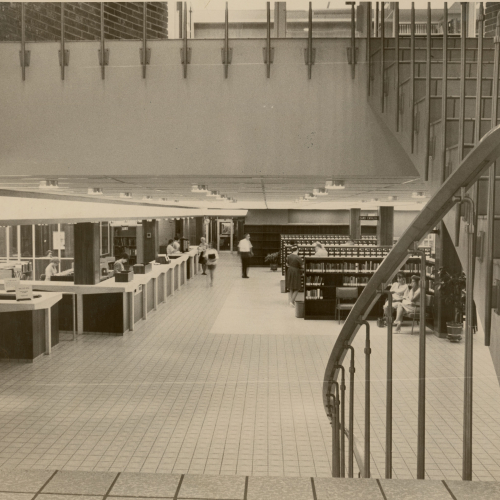 Taken from staircase, view toward main floor of library with service desk on left and stacks on right