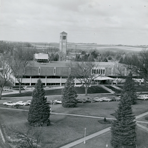 High-shot view of a brick and stone building with cars parked in front of it and Campanile and open space in the background