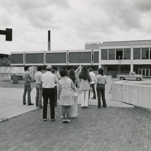People standing on roof of building looking toward brick and stone building with bank of windows on front