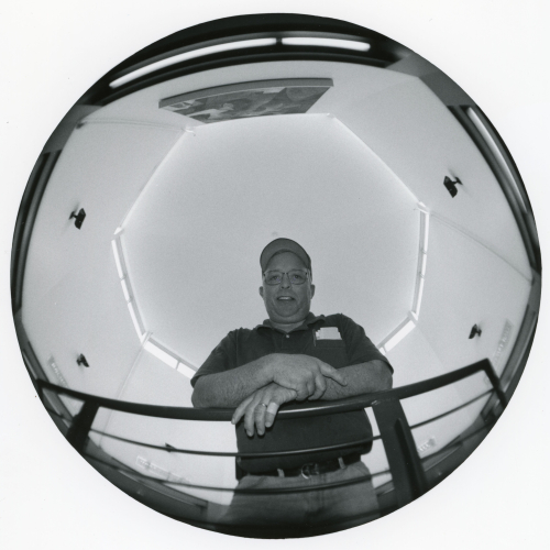 Fish-eye view of man in polo and hat leaning on rail, looking down into security camera
