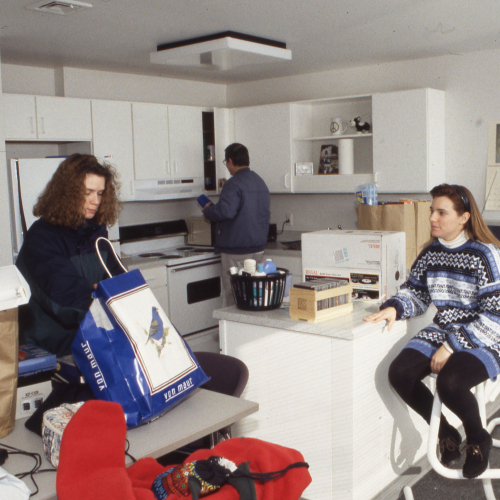 Two women unpacking in an apartment kitchen area, a man putting items into the cupboard behind them