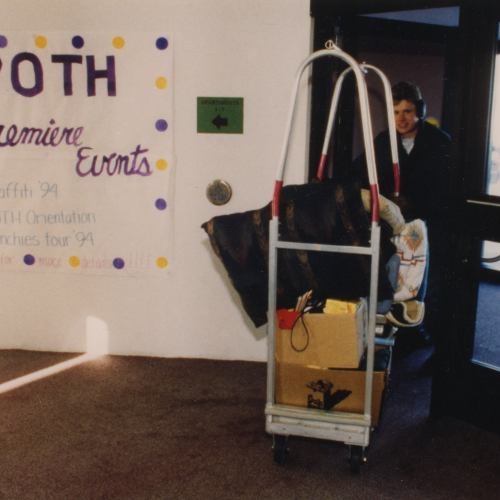 A person pushing a luggage trolley into a building; a poster on the wall reads: ROTH Premiere Events, Graffiti '94, ROTH Orientation, Munchies tour '94
