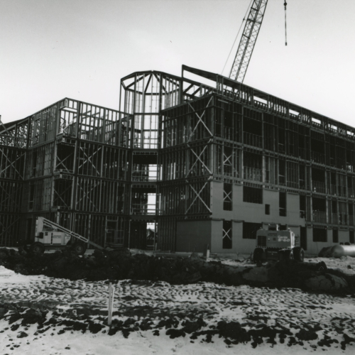 Construction in progress on building with two wings and tower framed in steel, some bricks placed along bottom levels of right wing