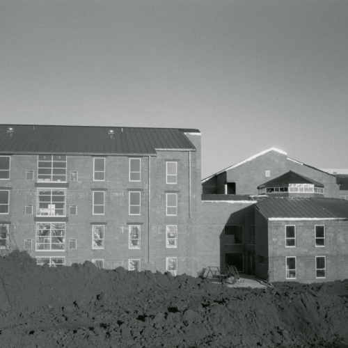 Mostly-completed four-story brick building with in-construction one behind it; windows still have plastic and dirt is not landscaped