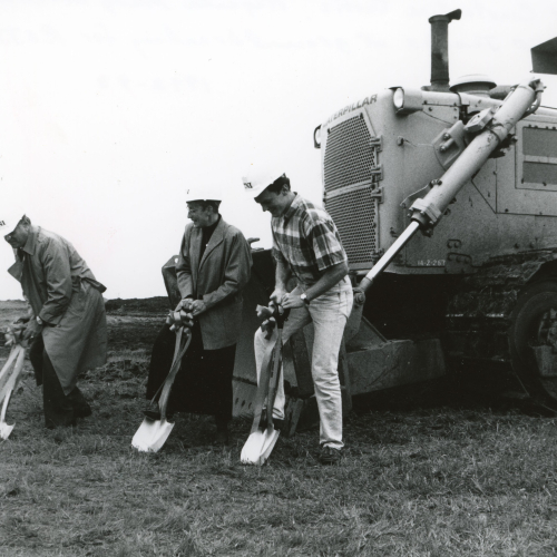 Three people in hard hats with shovels break ground, bulldozer behind them