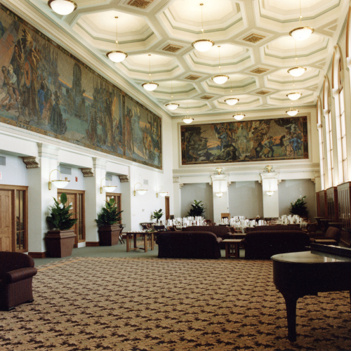Large room, half empty and half with tables set with dishes, murals on the upper portions of the left and back walls, windows on right