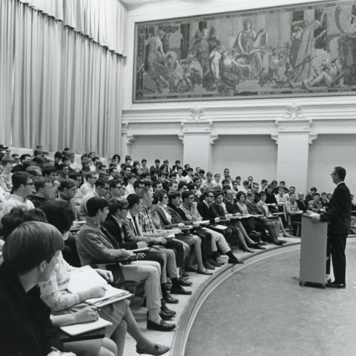 Professor lecturing to students filling auditorium-style seating, mural on wall behind