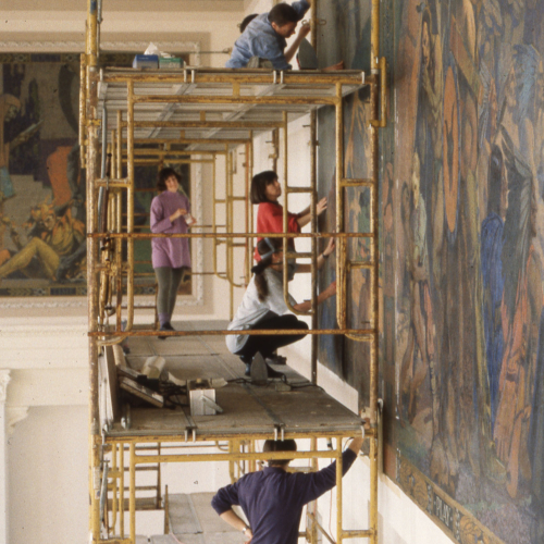 Six workers on three levels of scaffolding, working to put up mural on wall