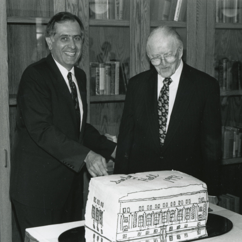 Two men cutting a cake shaped and decorated like a building, wording on top of cake reads Seerley Hall 1993