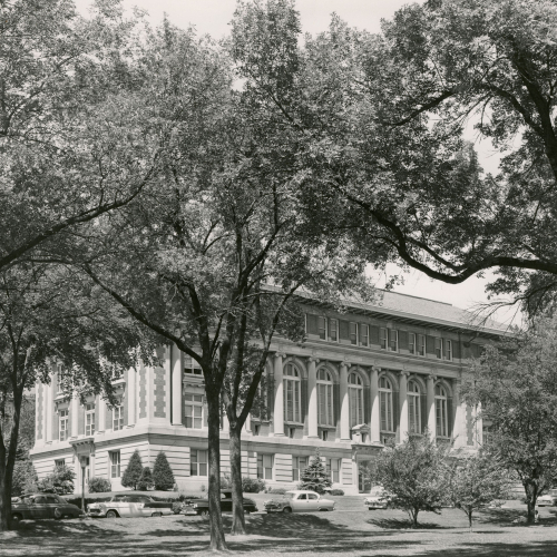 Framed by trees and with cars parked on street in front, a brick and limestone building with pillars and arched windows