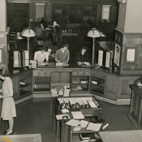 Woman working behind large library desk and three women filling out book information at the counter, library stacks visible beyond