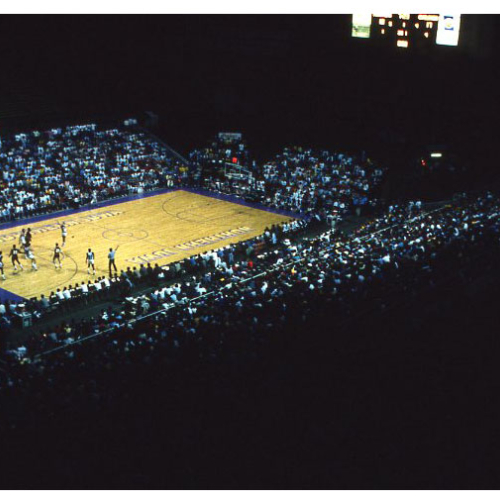 Shot from upper level, stadium seats dark around lighted basketball court and scoreboard