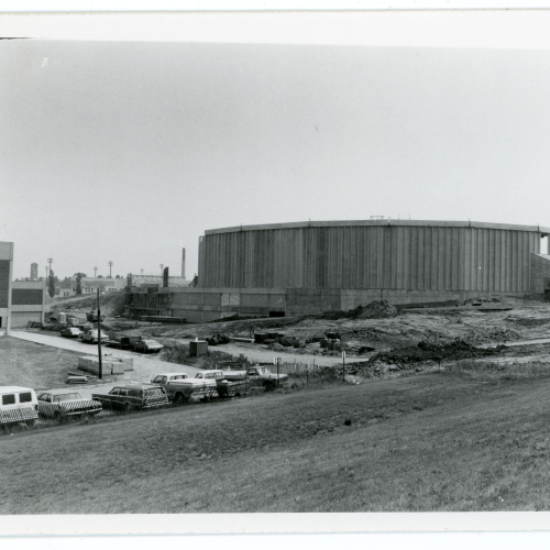 Crane next to partially-built walls of oval building, cars parked around cleared construction site