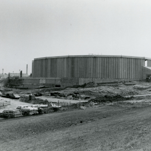 Crane next to partially-built walls of oval building, cars parked around cleared construction site