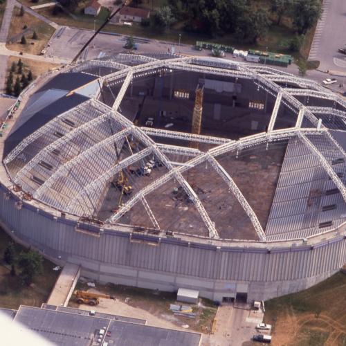 Roof beams with cables being installed on an oval-shaped building; interior of stadium is visible where roof panels haven't been installed