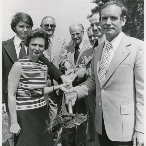 Seven people clustered around and holding a shovel with dirt on the blade