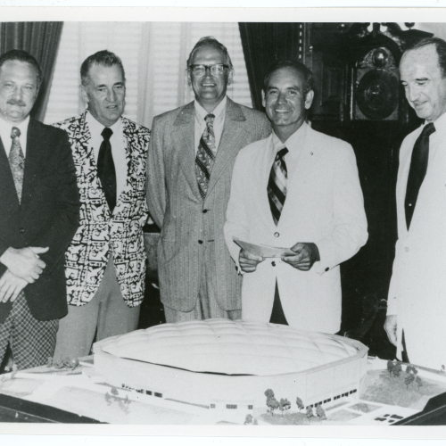 Five men standing behind a table which holds a model of an oval building with domed fabric roof