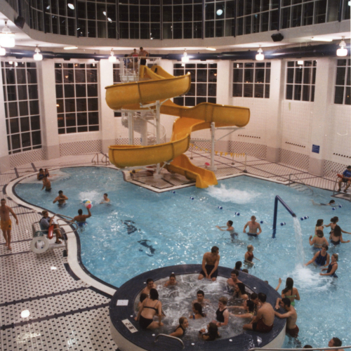 People in an indoor pool area with hot tub in foreground and yellow waterslide in background; the room has large windows and a vaulted ceiling
